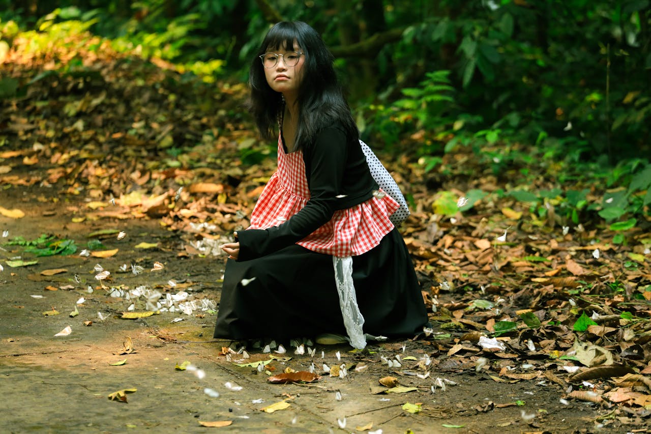 A young woman in a checkered red apron sitting on a leaf-strewn forest path in Ninh Bình, Vietnam.