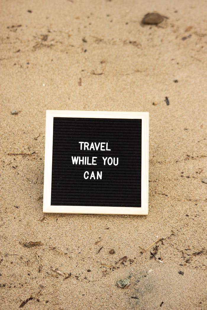 Inspirational message 'Travel while you can' on a signboard placed on beach sand, encouraging exploration.